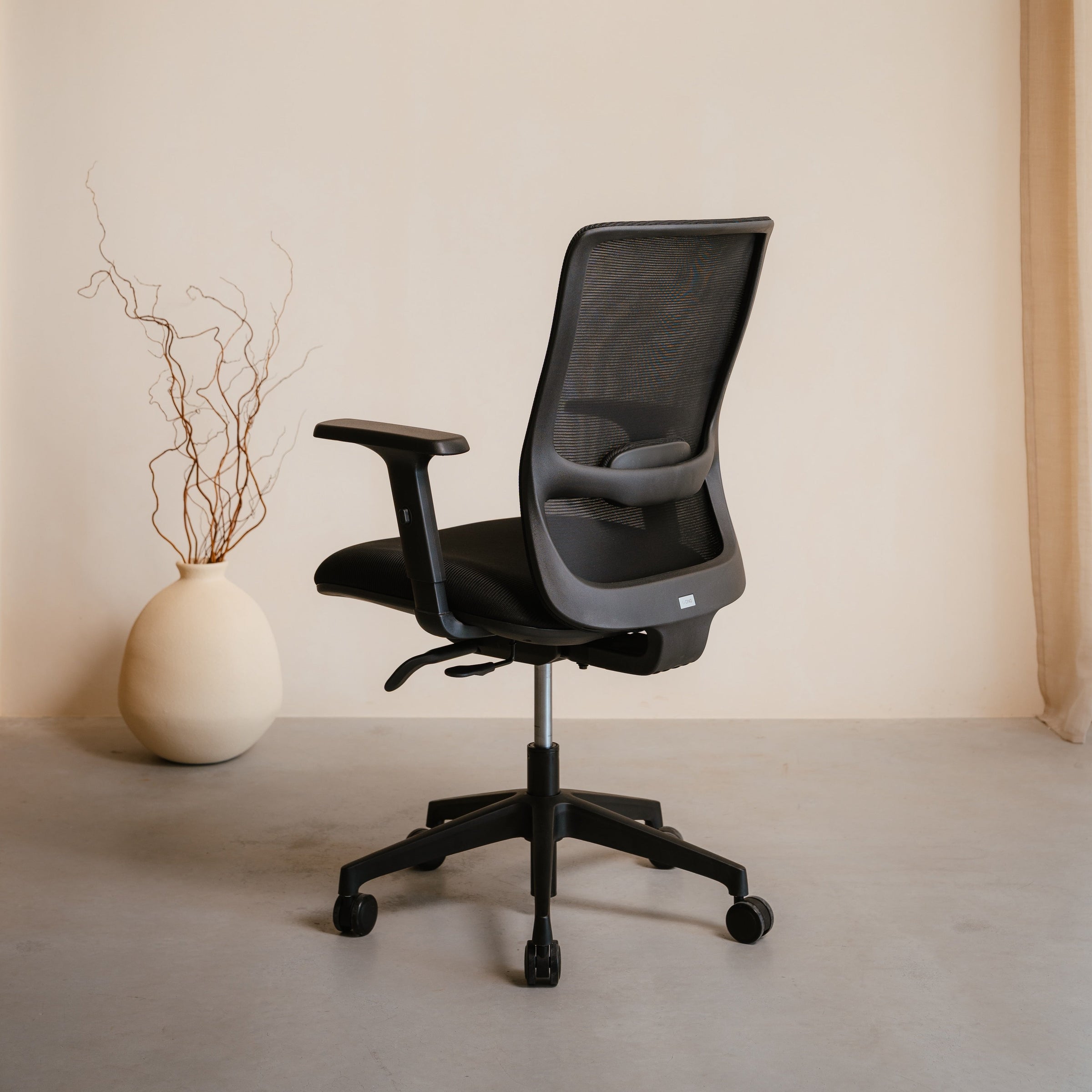 Black office chair in a minimalistic room with a vase and branches on a light-colored floor.